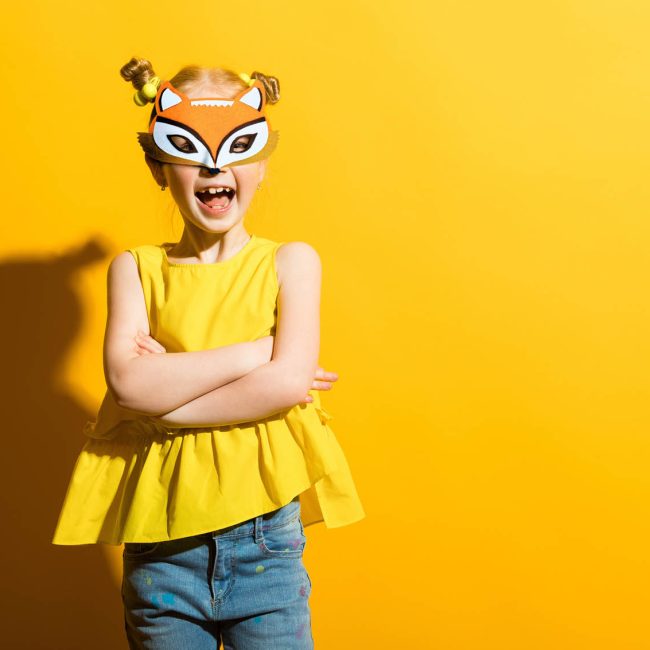 Petite fille avec couettes souriant, avec un masque renard sur le visage, sur fond jaune