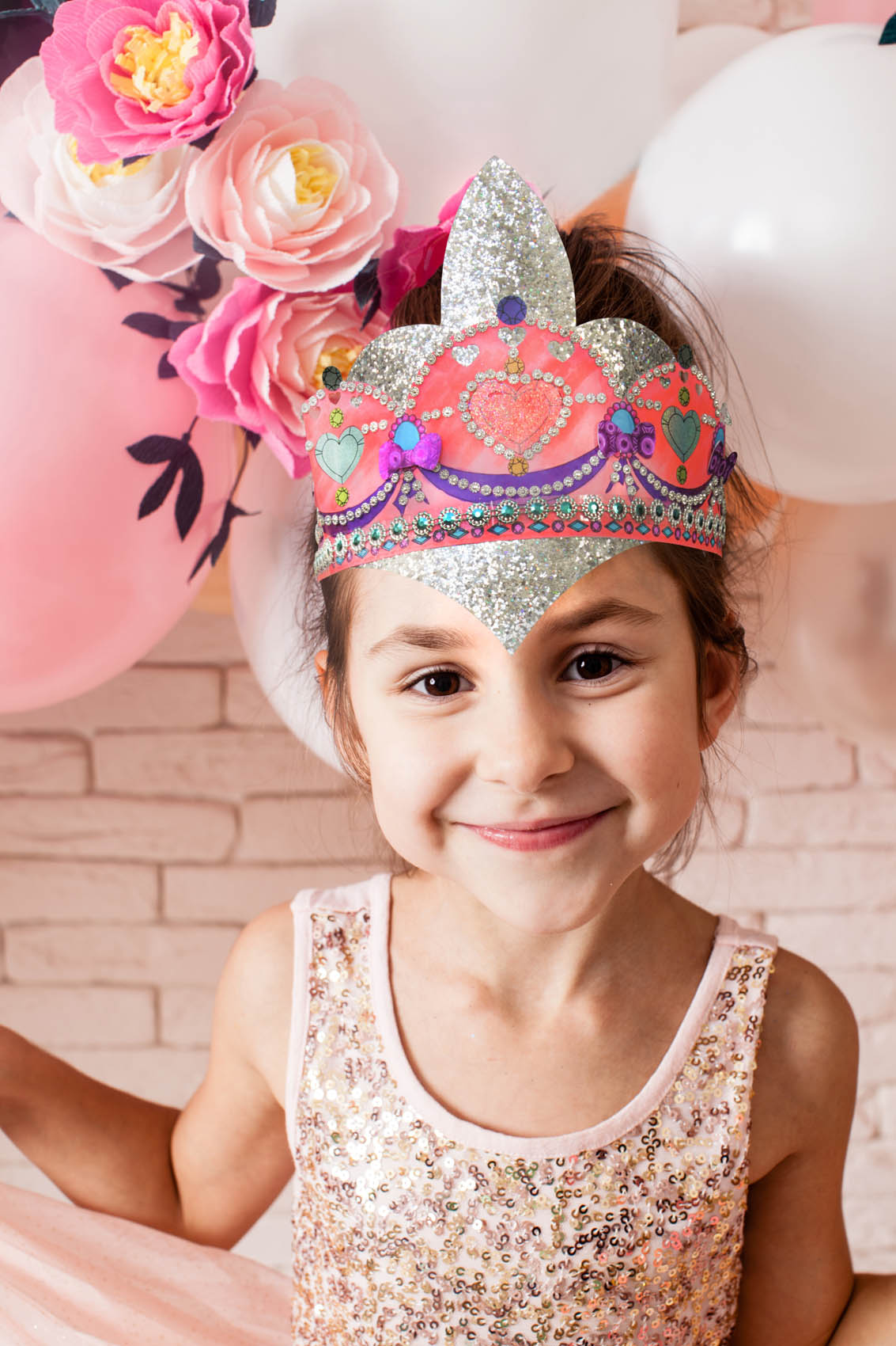 Petite fille en habit de fête souriant avec couronne décorée sur la tête et ballons derrière