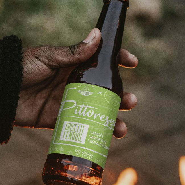 Bière avec étiquette verte tenue par une main noire, devant un feu de camp. Craft beer