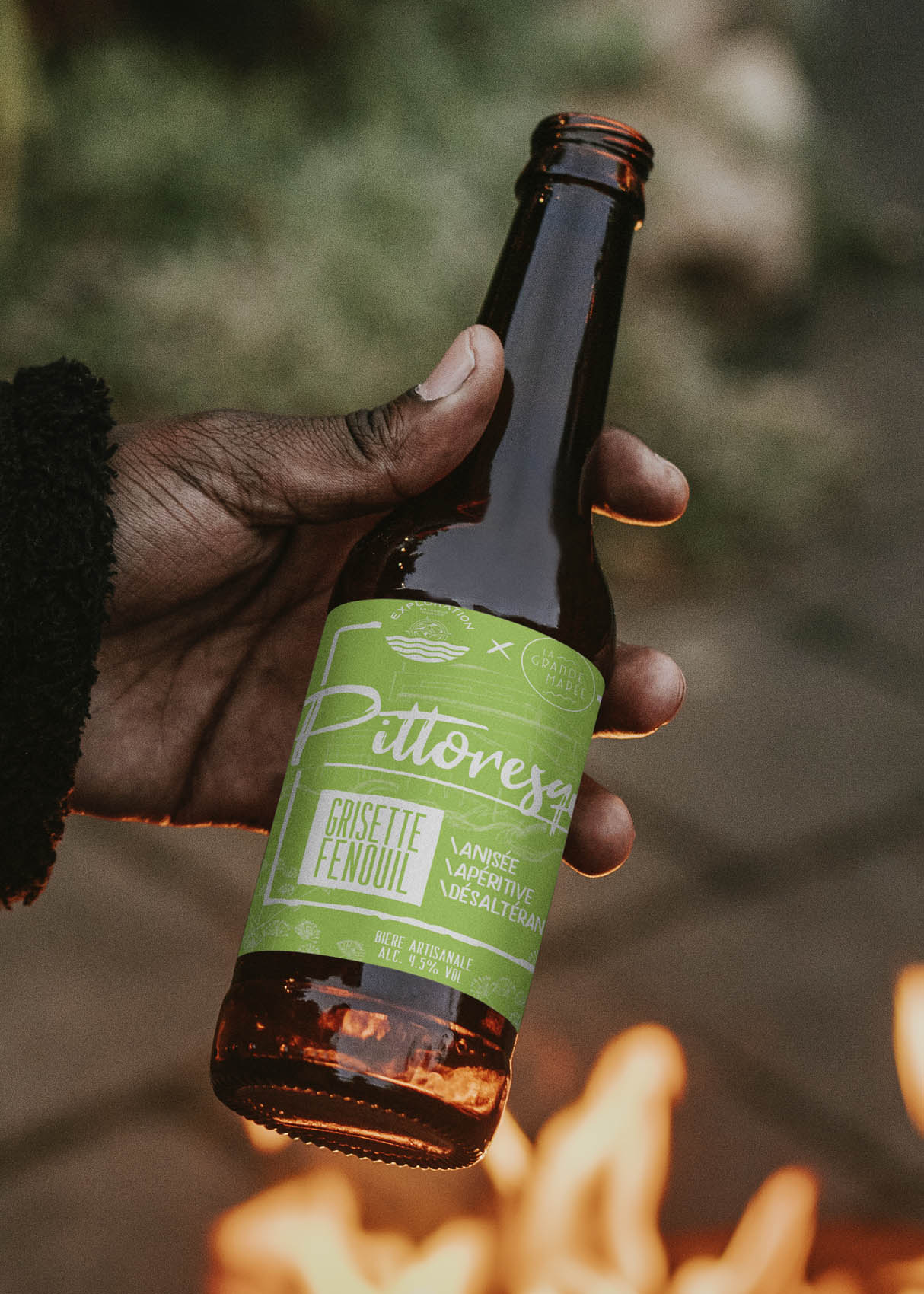 Bière avec étiquette verte tenue par une main noire, devant un feu de camp. Craft beer